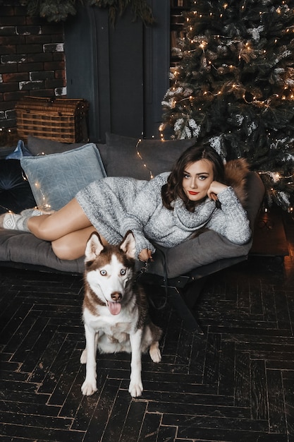 La mujer está sentando con husky en un sofá cerca del árbol de Navidad.