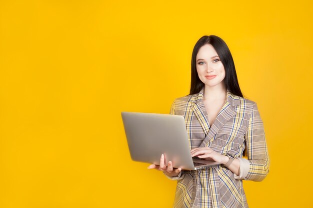 La mujer está sosteniendo una computadora portátil, con una sonrisa amistosa ligera, en un fondo amarillo, copie el espacio. El concepto de trabajar en una computadora y las nuevas tecnologías modernas.