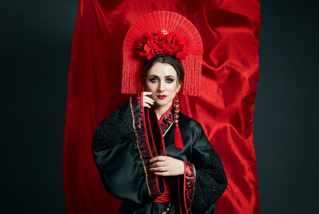 La mujer está vestida con ropa popular china japonesa roja. Tela voladora, bonito paraguas y abanico en estilo chino japonés, pendientes largos en las orejas. Chica posando sobre un fondo oscuro