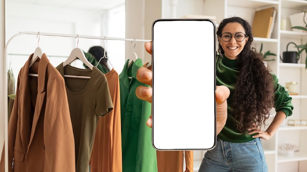 Mujer estilista de aplicaciones de moda que muestra un teléfono inteligente en blanco mientras está de pie cerca del riel de ropa