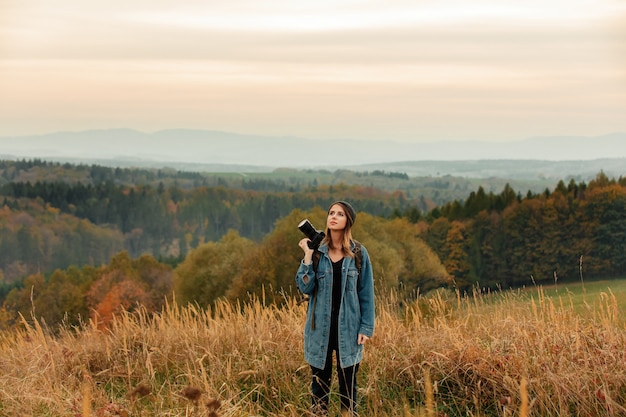 Mujer de estilo con cámara de fotos y mochila en el campo con montañas