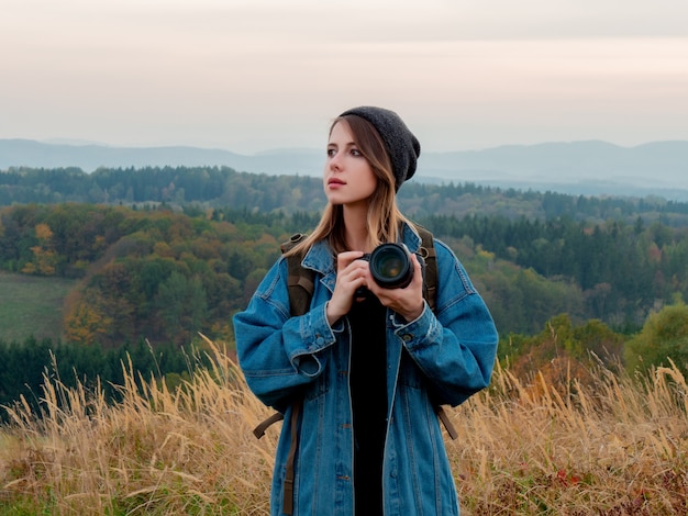 Mujer de estilo con cámara de fotos y mochila en el campo con montañas