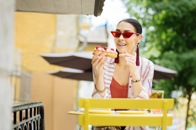 Mujer con estilo. Elegante mujer joven con gafas rojas y sonriendo mientras está sentado en un café y comiendo increíbles canutillos