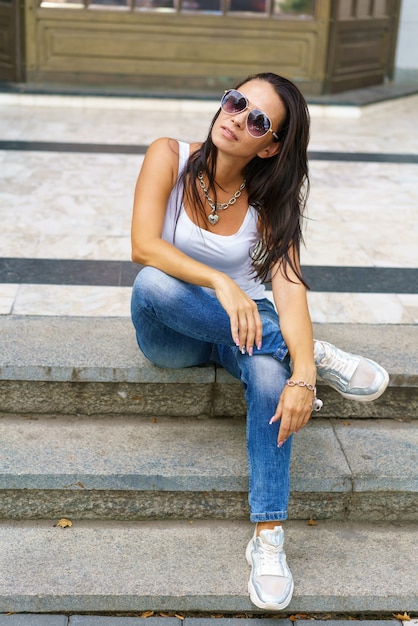 Mujer con estilo de estilo casual en gafas de sol sentado en una escalera de piedra que llevaba jeans un blanco ...