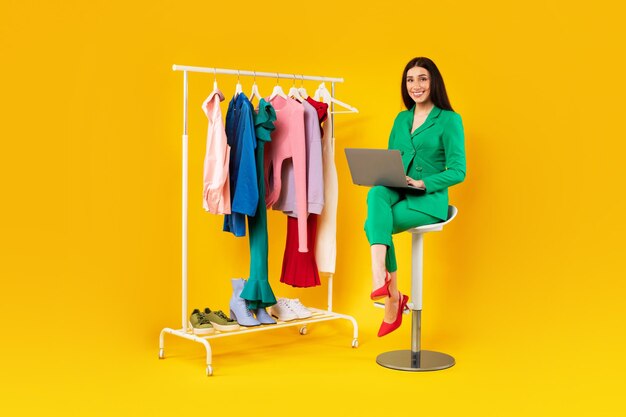 Mujer con estilo feliz usando una computadora portátil sentada en una silla cerca del riel de la ropa con ropa colorida en amarillo