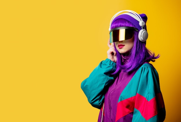 Mujer de estilo en gafas de realidad virtual y chándal de los años 80 con auriculares en la pared amarilla