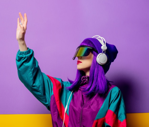 Mujer de estilo en gafas VR y chándal de los 80 con auriculares en la pared violeta