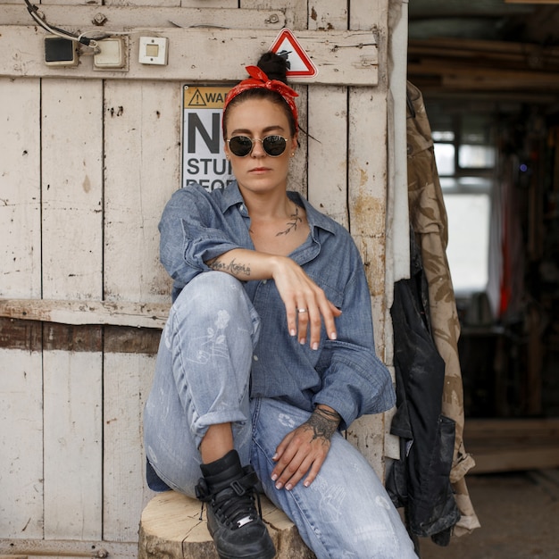 Foto mujer con estilo hipster con gafas de sol de moda en una camisa de mezclilla de moda y jeans sentado cerca de una pared de madera vintage