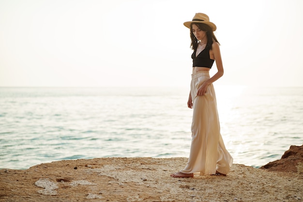 Mujer con estilo joven de pie sobre un acantilado en el mar al atardecer