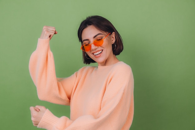 Mujer con estilo joven en suéter de melocotón casual y gafas naranjas aisladas en la pared de olivo verde positivo haciendo gesto ganador apretando espacio de copia de puño