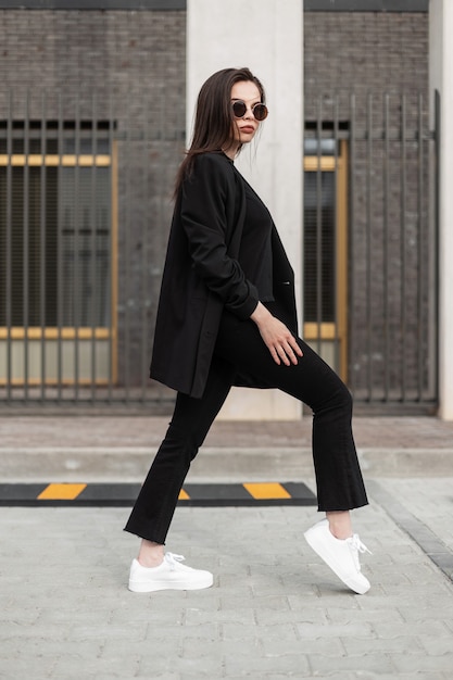 Mujer con estilo de moda en elegante blazer largo en pantalones de mezclilla vintage en camiseta cerca de edificio moderno en la calle. Chica de moda en ropa vintage en la ciudad. Traje de primavera de moda. Moda casual.