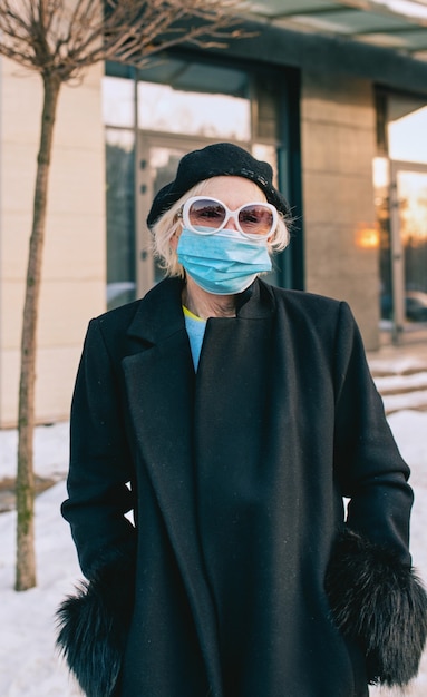 Mujer con estilo senior en boina y elegante abrigo negro y máscara médica caminando al aire libre