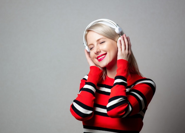 Mujer de estilo en suéter rojo con auriculares en pared gris