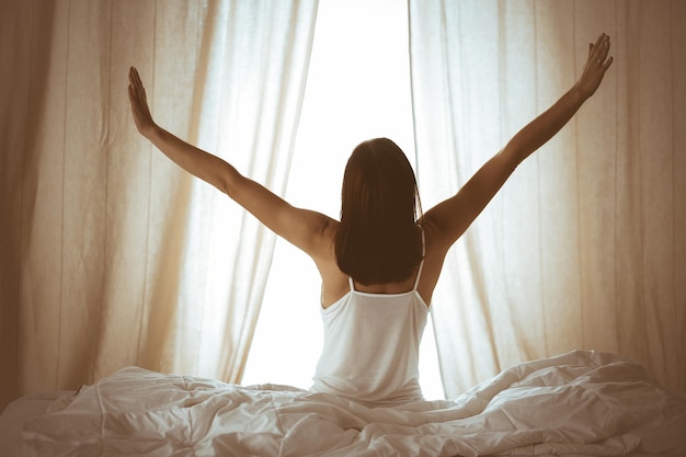 Mujer estirando las manos en la cama después de despertarse, llamarada solar. Morena entrando en un día feliz y relajado después de una buena noche de sueño y vista trasera. Concepto de un nuevo día y un fin de semana alegre.