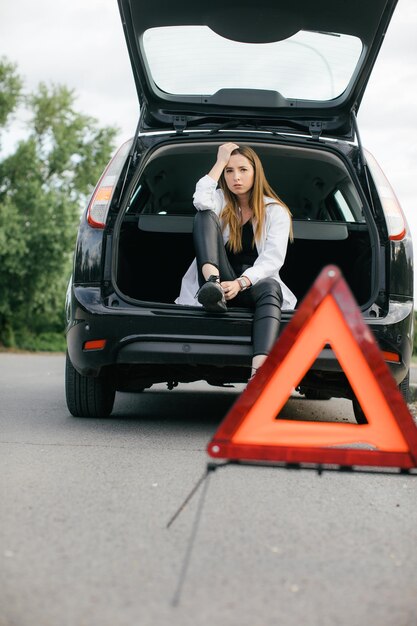 Mujer estresada mirando un auto roto y revisando el motor en la carretera rural a la vista lateral