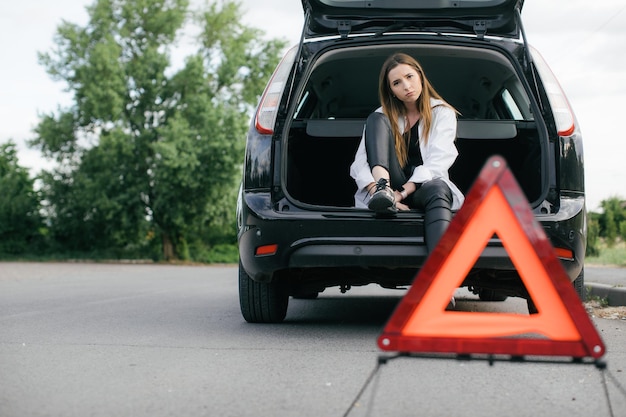 Mujer estresada mirando un auto roto y revisando el motor en la carretera rural a la vista lateral