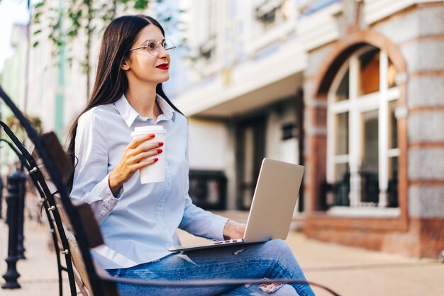 Mujer estudiante bastante sonriente vestida informalmente sentada al aire libre en un banco disfrutando de su café y trabajando con ordenador portátil