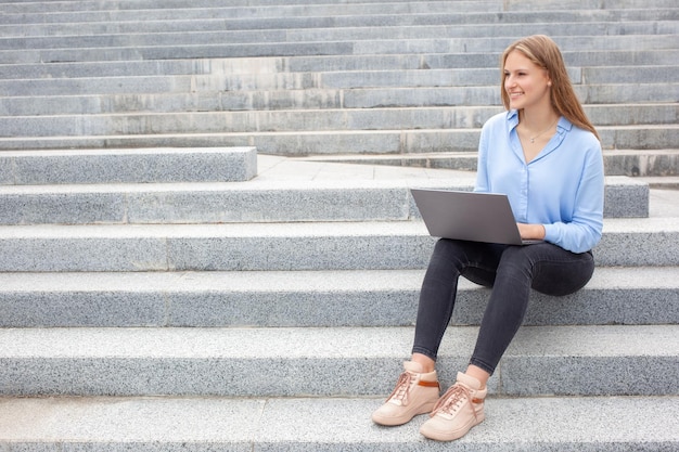 Mujer estudiante europea con cara feliz sentada en las escaleras y trabajando en una laptop mirando hacia otro lado