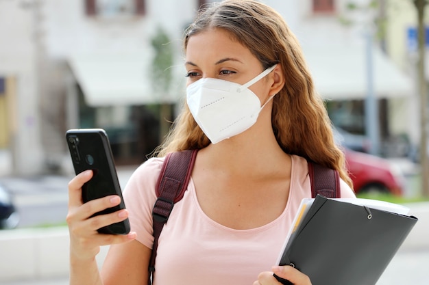 Mujer estudiante con máscara protectora leyendo un mensaje en un teléfono inteligente en la calle