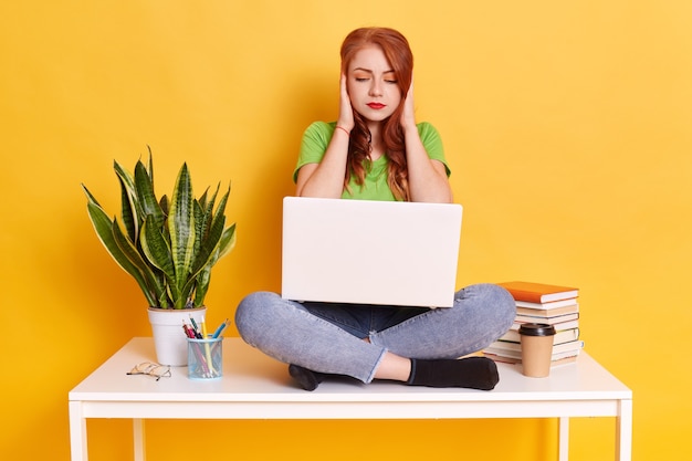 Mujer estudiante sentada en la mesa con el portátil sobre las rodillas, cubriendo los oídos con las palmas, ignorando el molesto ruido