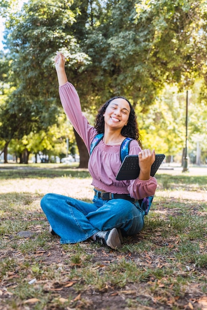 Mujer estudiante universitaria latina celebrando y sosteniendo una tableta sentada afuera en un parque en el césped