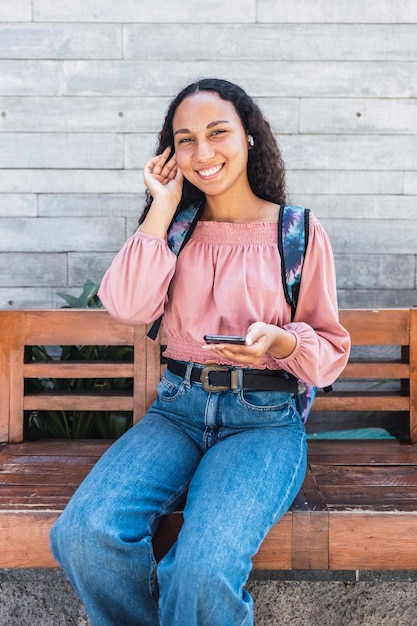 Mujer estudiante universitaria latina sonriendo y usando su móvil sentada afuera del centro comercial en tiempo libre