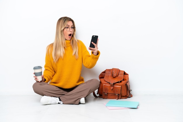 Mujer estudiante uruguaya sentada en el suelo aislada de fondo blanco sosteniendo café para llevar y un móvil