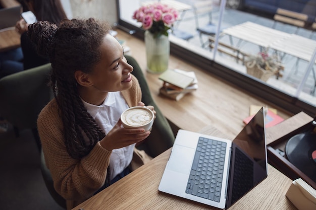 Mujer étnica confiada con una taza de café mirando soñadoramente por la ventana a la calle de la ciudad mientras