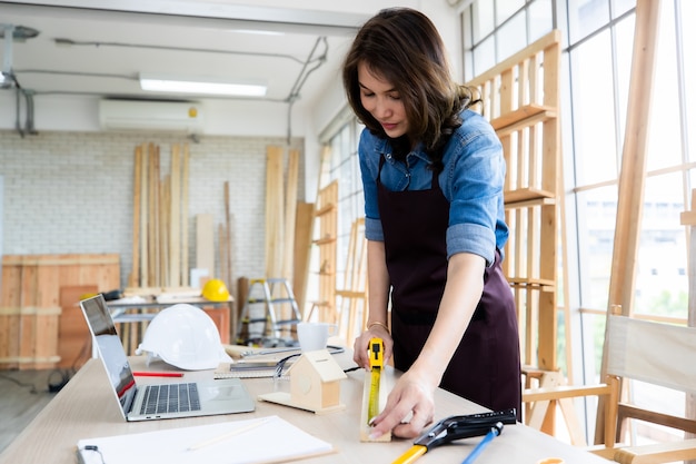 Mujer étnica positiva en delantal sonriendo y midiendo el tablero de madera en la mesa cerca de la computadora portátil mientras trabaja en carpintería ligera