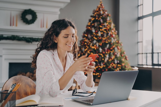 Mujer europea alegre sentada en el escritorio con una computadora portátil leyendo noticias de Navidad o un mensaje en el teléfono inteligente