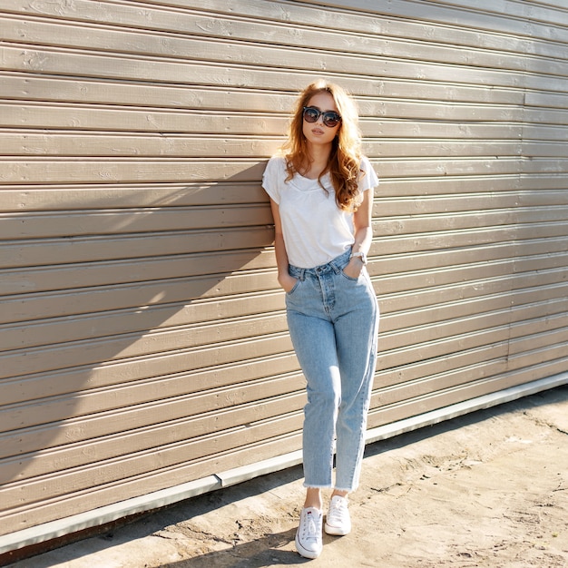 La mujer europea bastante joven inconformista en elegante camiseta blanca en jeans vintage con gafas de sol de moda está de pie cerca de la pared de madera y disfruta del verano
