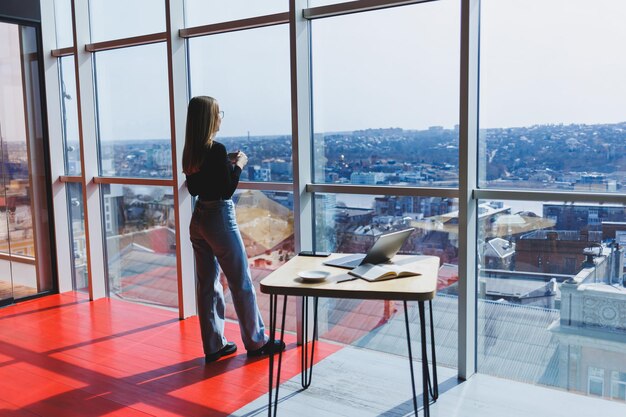 Mujer europea bebe café y mira por la ventana en un café Una joven sonriente con gafas se para cerca de la ventana Ocio recreativo y tiempo libre Estilo de vida femenino moderno