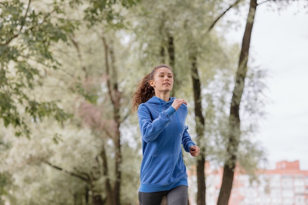 Una mujer europea con una capucha azul corre y hace deporte en el parque, fitness y yoga, deportes al aire libre