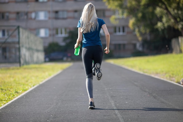 Mujer europea delgada corriendo en el estadio Chica alegre en pantalones negros sosteniendo una botella de agua durante el entrenamiento al aire libre.