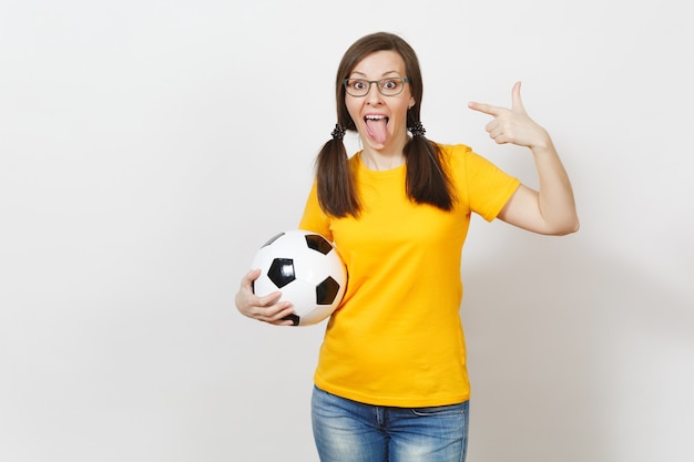 Mujer europea, dos divertidas colas de caballo, aficionado al fútbol o jugador con gafas, uniforme amarillo mostrar lengua mantenga clásico balón de fútbol aislado sobre fondo blanco. Deporte jugar al fútbol, concepto de estilo de vida saludable.