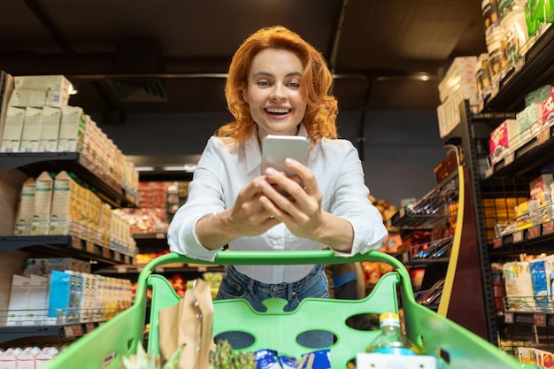 Mujer europea feliz haciendo compras de comestibles y usando teléfono móvil posando con el carrito del tranvía en