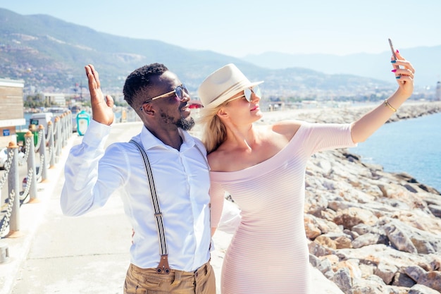 Mujer europea feliz sonriente con sombrero de paja y vestido rosa sexy haciendo un retrato selfie en la cámara del teléfono inteligente con un hombre étnico afroamericano en la playa de verano con vistas al rock en el complejo tropical de Turquía.