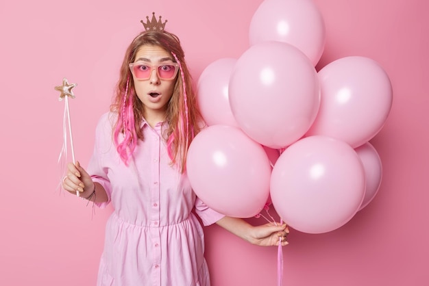 Una mujer europea impresionada y atónita usa gafas de sol de moda y el vestido reacciona ante impactantes noticieros montón de globos inflados y varita mágica aislado sobre fondo rosa Concepto de evento de vacaciones