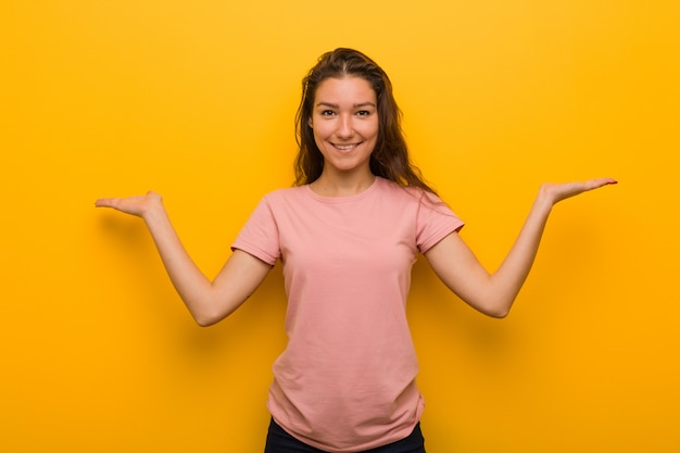 La mujer europea joven aislada sobre amarillo hace escala con los brazos, se siente feliz y confiada.