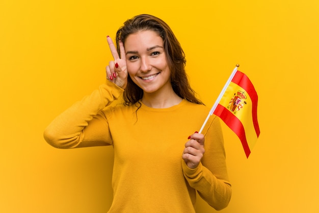 Foto mujer europea joven que sostiene una bandera española que muestra la muestra de la victoria y que sonríe ampliamente.