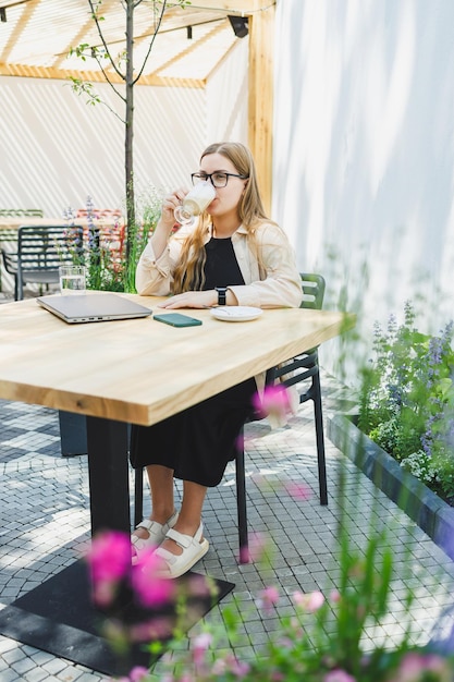 Mujer europea joven sentada en una mesa de café al aire libre con una computadora portátil y una taza de café mujer sonriente con gafas disfrutando del teletrabajo en una cafetería o estudiando en línea