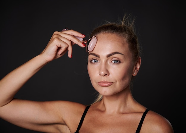 Mujer europea de mediana edad con piel bronceada brillante y saludable masajeando la cara con rodillo de jade aislado sobre fondo negro con espacio publicitario para copiar Terapia de belleza de rejuvenecimiento suavizante antienvejecimiento