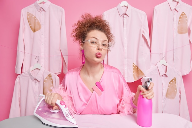 La mujer europea ocupada de pelo rizado mantiene los labios doblados ocupados haciendo planchado usa agua en aerosol usa lentes transparentes bata trabaja en lavandería tiene expresión romántica. Concepto de limpieza.