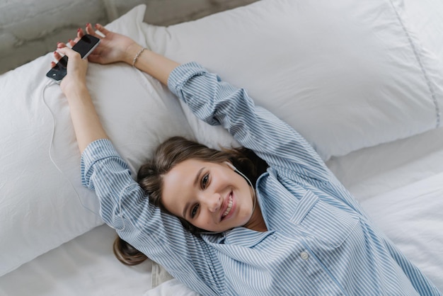 Una mujer europea relajada con dientes disfruta de un buen descanso en la cama se estira durante la mañana sostiene un teléfono celular moderno escucha su canción favorita con auriculares se regocija de comenzar un nuevo día Vista horizontal