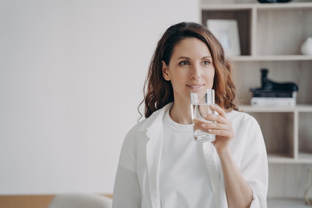 Mujer europea relajada está bebiendo agua pura en casa Chica feliz está sosteniendo un vaso y sonriendo