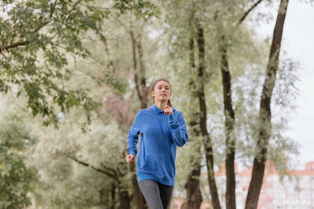 Una mujer europea con una sudadera con capucha azul corre y hace deporte en el parque, fitness y yoga al aire libre