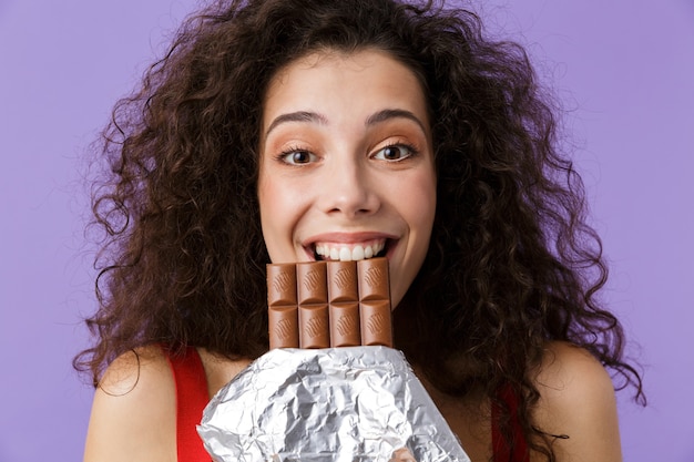 Mujer europea con vestido rojo sonriendo y comiendo chocolate, que se encuentran aisladas sobre la pared violeta