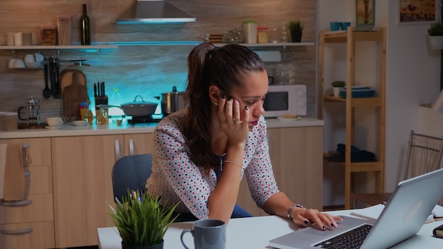 Mujer con exceso de trabajo quedarse dormido en la cocina de casa a altas horas de la noche. Empleado remoto agotado ocupado tomando una siesta en una silla despertando trabajando en una computadora portátil usando la red de tecnología moderna inalámbrica haciendo horas extras.