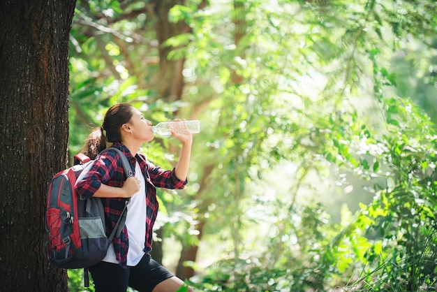 Mujer excursionista bebiendo agua mientras se apoya en el tronco de un árbol en el bosque