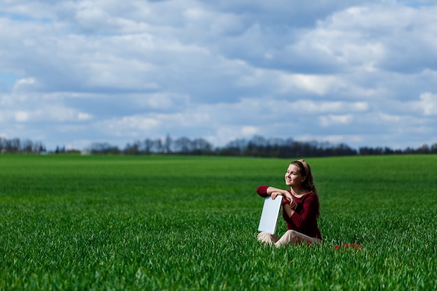 Mujer de éxito joven está sentada sobre la hierba verde con una computadora portátil en sus manos. Descanse después de una buena jornada laboral. Trabaja en la naturaleza. Chica estudiante trabajando en un lugar apartado. Lugar de trabajo en la naturaleza